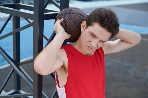 Ein junger Mann im roten Hemd hält gekonnt einen Basketball in der Hand, während er sich darauf vorbereitet, an einem sonnigen Sommertag im Freien zu spielen.. — Stockfoto
