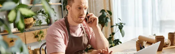 Un uomo in un negozio di piante, seduto a un tavolo, impegnato in una telefonata. — Foto stock