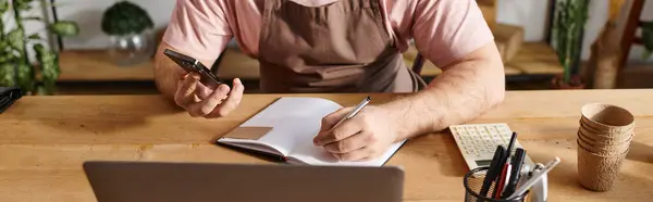 Ein Mann mitten in der Arbeit, der mit Notizbuch und Handy an einem Schreibtisch sitzt und sich auf seine kleinen Geschäftsideen konzentriert. — Stockfoto