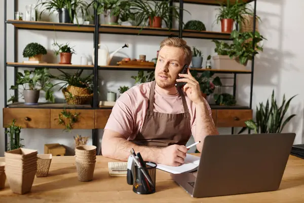 Un hombre se sienta en una mesa en una tienda de plantas, hablando por un teléfono celular mientras maneja su pequeño negocio. - foto de stock