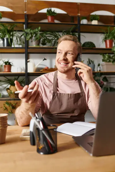 Un homme assis à une table dans un magasin de plantes, parlant sur un téléphone portable tout en étant entouré de verdure. — Photo de stock