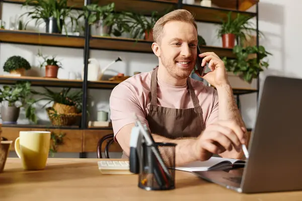 Un hombre muy ocupado trabaja en su computadora portátil en una tienda de plantas mientras tiene una conversación telefónica. - foto de stock