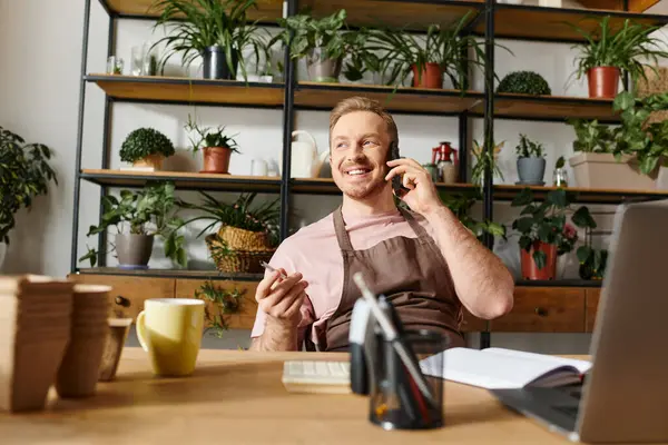 Un hombre absorto en la conversación en un teléfono celular, sentado en un escritorio en una bulliciosa tienda de plantas. - foto de stock