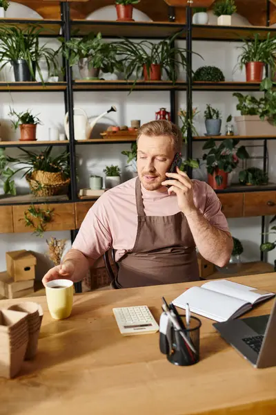 A man sits at a desk in a plant shop, talking on a cell phone, showcasing determination towards his small business success. — Stock Photo
