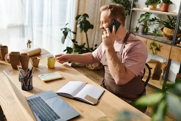 Um homem bonito sentado em uma mesa em uma loja de plantas, profundamente envolvido em uma conversa em seu telefone celular. — Fotografia de Stock