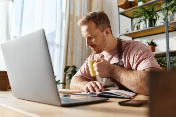Un homme assis sur un ordinateur portable dans un espace de travail rempli d'usines, se concentrant sur ses opérations de petite entreprise. — Photo de stock