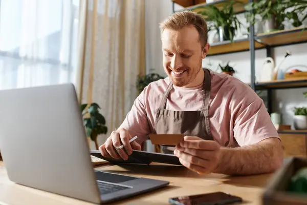 A man sitting at a laptop computer, holding a credit card, likely purchasing supplies for his small plant shop business. — Stock Photo