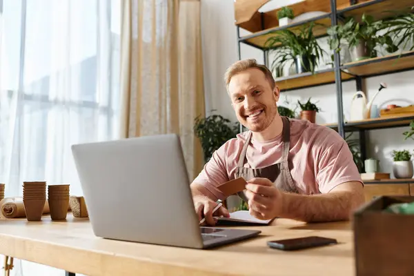 Um homem com um laptop e cartão de crédito, focado em seu pequeno negócio em uma loja de plantas. — Fotografia de Stock