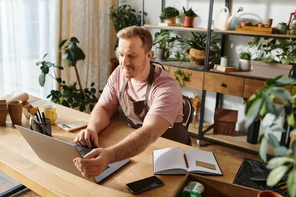 Um homem com um toque na moda senta-se em uma mesa, focado em seu laptop em meio a uma variedade exuberante de plantas em uma loja de plantas caseiro. — Fotografia de Stock