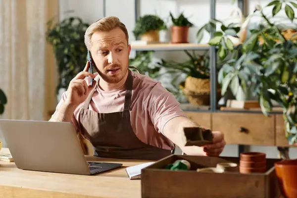 Un hombre se sienta en una mesa en una tienda de plantas, se dedica a una llamada telefónica mientras atiende a su pequeña empresa. - foto de stock