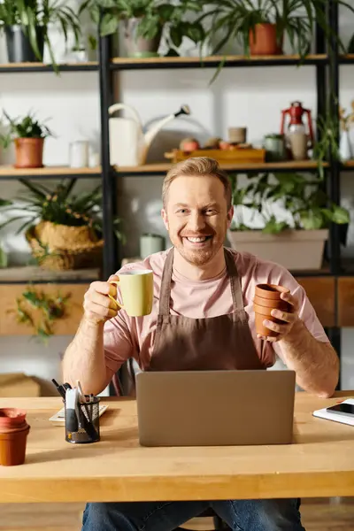 Ein Mann sitzt neben seinem Laptop und hält Kaffee und Kanne an einem Tisch in einem Pflanzenladen. — Stockfoto
