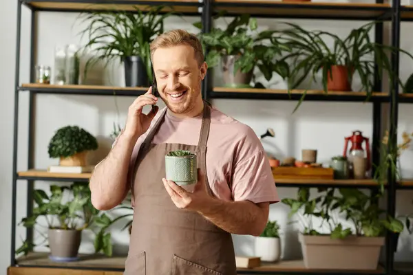 A man in an apron multitasks by holding a plant pot and talking on a cell phone in a botanical shop. — Stock Photo
