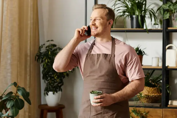 Ein gutaussehender Mann in einer Schürze eines Pflanzengeschäfts, der mit einem Handy spricht und Kleinunternehmer in der Blumenindustrie präsentiert. — Stockfoto