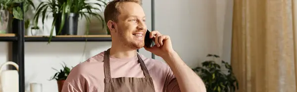 Un hombre con estilo en un delantal conversa en un teléfono celular, administrando su tienda de plantas con un enfoque en el servicio al cliente. - foto de stock