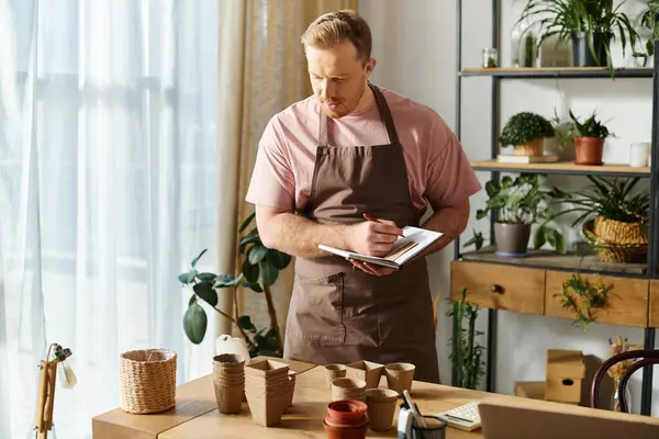 A man in an apron diligently takes notes on a clipboard in a plant shop, showcasing his dedication to his small business. — Stock Photo