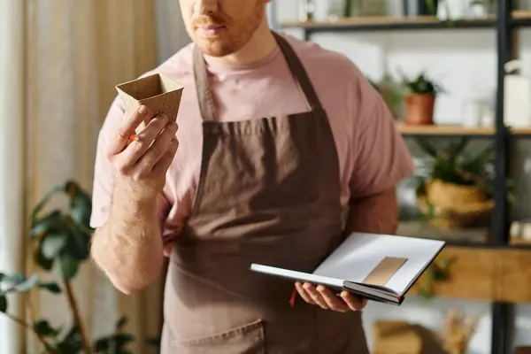 A handsome man in an apron standing with a notebookin a plant shop, showcasing a sense of ownership and dedication. — Stock Photo