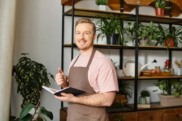 A handsome man in an apron holds a notebook, managing his plant shop with a business owners dedication. — Stock Photo