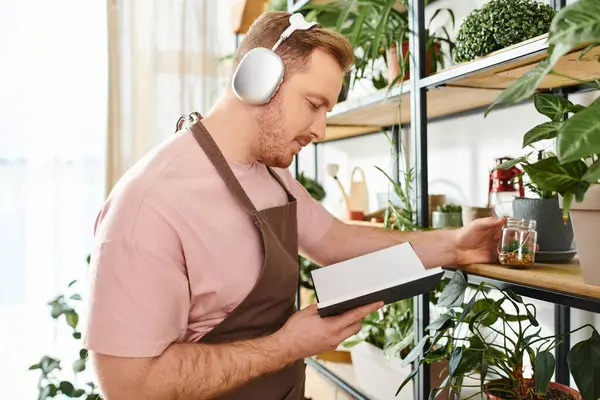 Um homem usando fones de ouvido está atentamente olhando para um caderno em uma loja de plantas, refletindo sobre a beleza da natureza.. — Fotografia de Stock