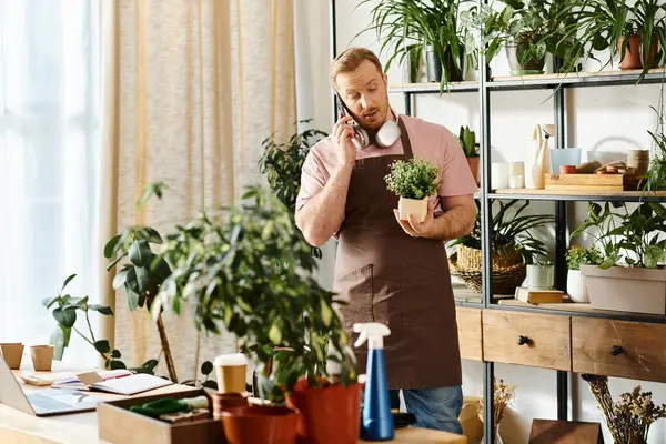 Um homem em um avental fala em um telefone celular enquanto mantém uma planta em vaso em uma loja de plantas. — Fotografia de Stock