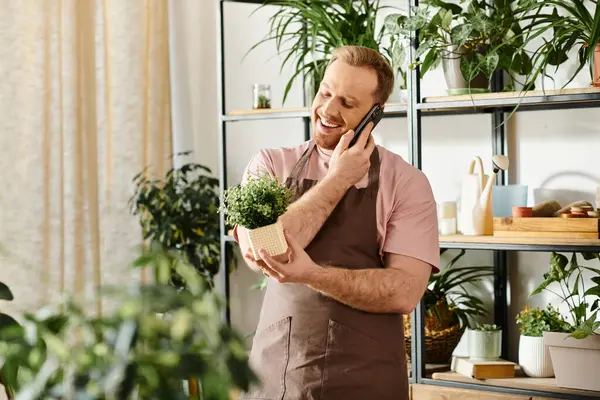 Um homem elegante multitarefas, conversando em um celular enquanto delicadamente segurando uma planta em vaso em uma loja de plantas. — Fotografia de Stock