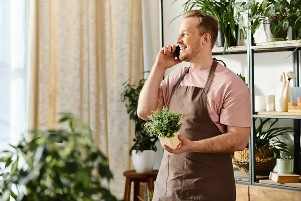A man chatting on cell phone and holding a potted plant in a plant shop, embodying multitasking and business ownership. — Stock Photo