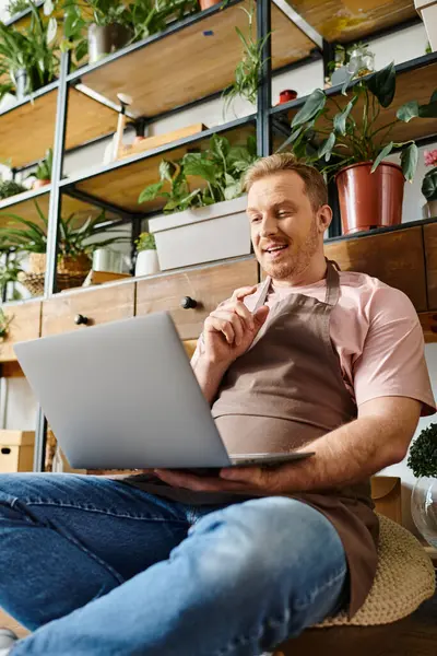 Um homem com um computador portátil senta-se no chão de uma loja de plantas, imerso no seu trabalho e rodeado por vegetação. — Fotografia de Stock