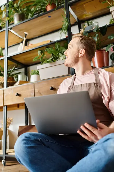 A man sitting on the floor, deeply focused on his laptop, surrounded by lush green plants in a plant shop. — Stock Photo