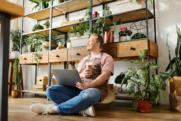 Um homem elegante se senta no chão, focado em seu laptop em sua pequena loja de plantas. Ele está ocupado gerenciando seu negócio florista. — Fotografia de Stock