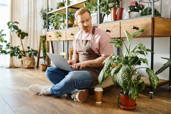 Un uomo in un negozio di piante si siede sul pavimento lavorando intensamente sul suo computer portatile, incarnando dedizione alla sua piccola impresa.. — Foto stock