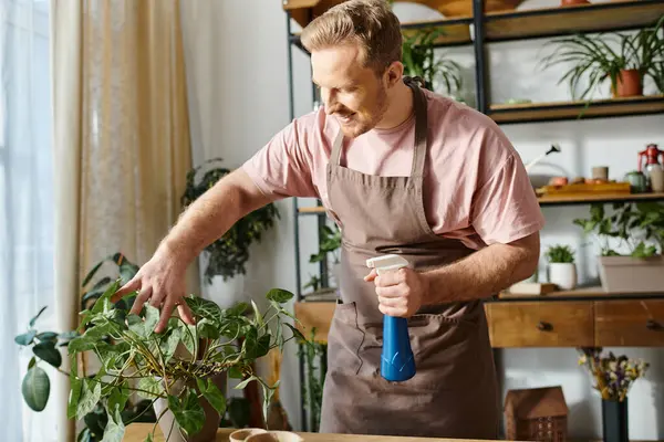 Un hombre en un delantal sostiene cuidadosamente una botella de spray azul en un entorno de tienda de plantas, mostrando su dedicación a su propio negocio. - foto de stock