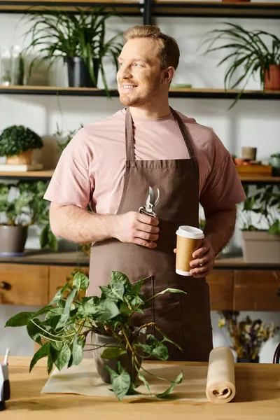 A man in an apron holding a cup of coffee in a plant shop. — Stock Photo