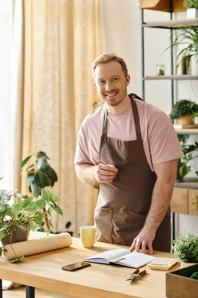 Um homem em um avental fica confiante na frente de uma mesa, mostrando suas habilidades como proprietário de uma loja de plantas. — Fotografia de Stock