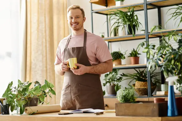 Um homem bonito em um avental desfruta de uma xícara de café em uma loja de plantas, incorporando a essência de possuir uma pequena empresa. — Fotografia de Stock