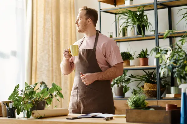 A handsome man in an apron enjoys a cup of coffee in a plant shop, embodying the essence of a small business owner. — Stock Photo