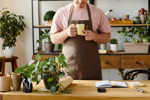 Um homem em um avental desfrutando de uma xícara de café em um ambiente de loja de plantas, tomando um momento para relaxar e saborear a bebida quente. — Fotografia de Stock