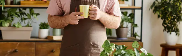 Hombre cortado pacíficamente sosteniendo una taza en sus manos mientras atiende a las plantas en su pequeña tienda de plantas. - foto de stock