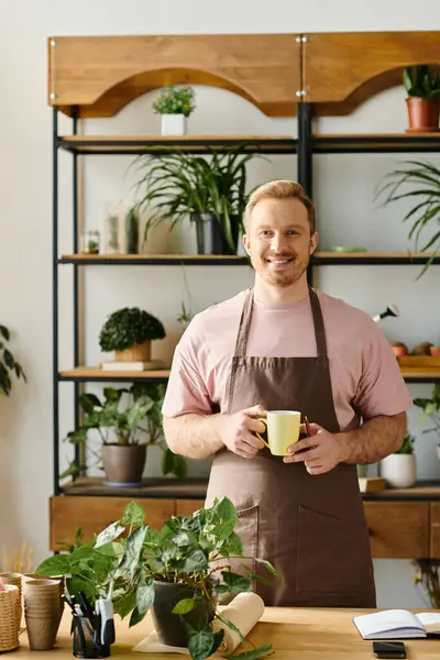Un hombre con estilo en un delantal disfrutando de una taza de café en una tienda de plantas, encarnando el propio concepto de negocio. - foto de stock
