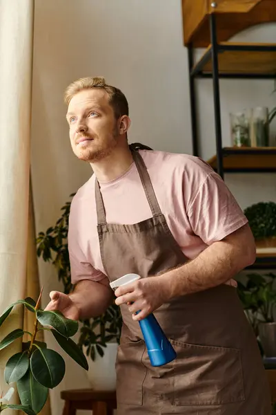 Um homem bonito em um avental segurando uma garrafa de spray azul em uma loja de plantas, refletindo o encanto de possuir um pequeno negócio floral. — Fotografia de Stock