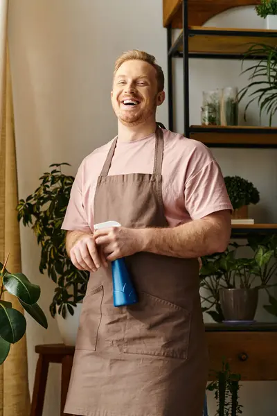 Un hombre en un delantal sostiene una botella de spray azul en una tienda de plantas, mostrando sus habilidades y pasión por su pequeña empresa. - foto de stock