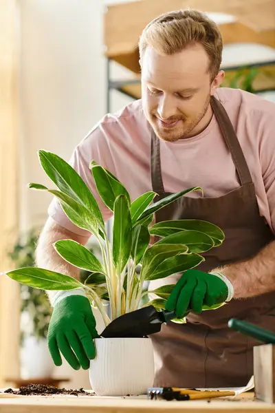 Un hombre con delantal y guantes atiende cuidadosamente a una planta en maceta en una pequeña tienda de plantas, encarnando la esencia de poseer un negocio floral. - foto de stock