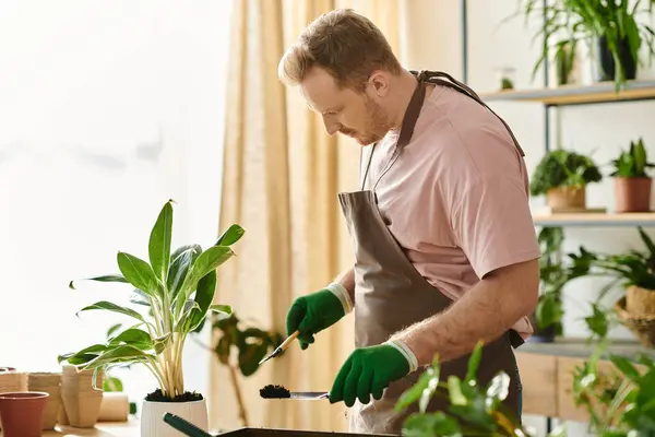 Un hombre con un delantal y guantes verdes prepara expertamente una planta en maceta en un encantador entorno de tienda de plantas. - foto de stock