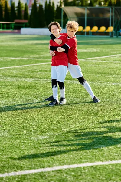 Deux jeunes garçons, vêtus d'équipement de football, s'embrassent amoureusement sur le terrain de football vert. Leurs visages rayonnent de bonheur et de sportivité alors qu'ils célèbrent ensemble. — Photo de stock