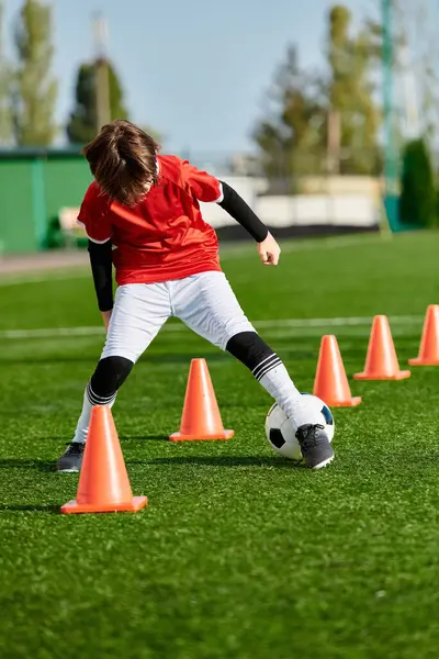 Талантливый мальчик умело маневрирует футбольным мячом вокруг ярких оранжевых конусов на поле, демонстрируя свою ловкость и точность в дриблинге и пинках.. — стоковое фото