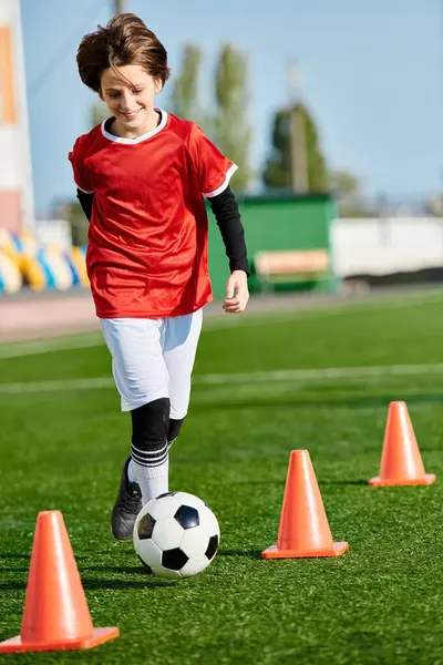 Un jeune garçon démontrant ses talents de footballeur en frappant un ballon de football autour de cônes orange sur un terrain. Ses pas précis et son agilité sont évidents alors qu'il navigue à travers les obstacles avec facilité. — Photo de stock