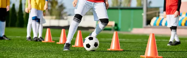 Un jugador de fútbol experto está pateando una pelota de fútbol alrededor de conos de color naranja en un campo. Demuestra técnicas precisas de goteo mientras navega a través de los obstáculos con agilidad y control. - foto de stock