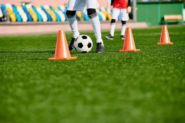Geschickt kickt ein gelernter Fußballer einen Fußball durch eine Reihe orangefarbener Kegel, die in einer Trainingsübung aufgestellt sind. Die Spieler sind fokussiert, wendig und beherrschen den Kurs mit Finesse. — Stockfoto