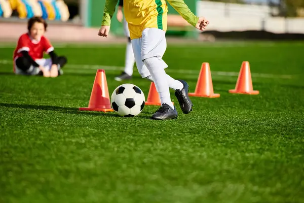 A determined young boy practices dribbling a soccer ball around cones on a sports field, showcasing his agility and precision in each kick. The vibrant energy of his movements captures the essence of youthful enthusiasm and athletic spirit. — Stock Photo