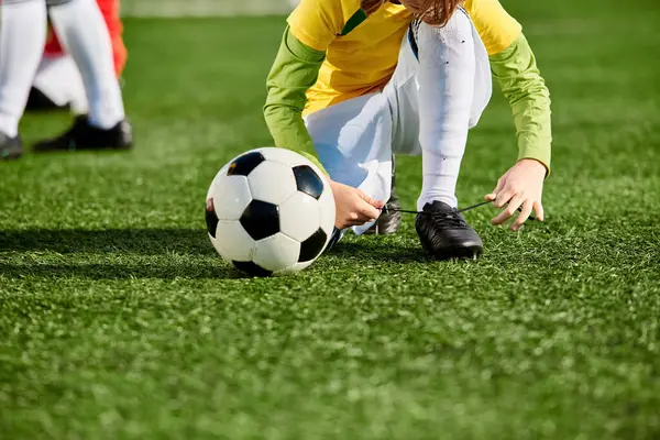 Una joven con coletas se arrodilla en un campo, alcanzando a recoger una pelota de fútbol con patrones de colores en ella. - foto de stock