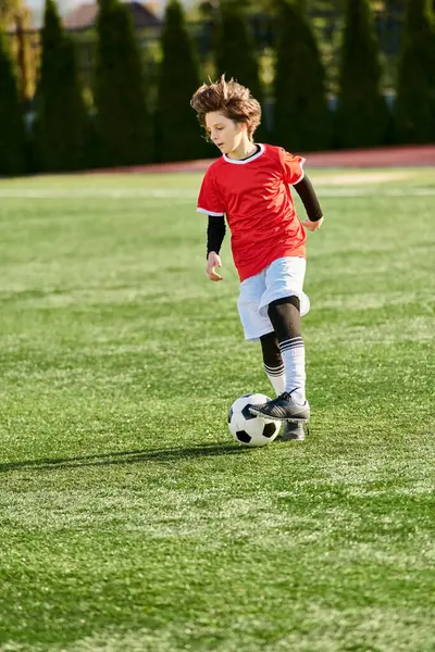 Молодой мальчик уверенно пинает футбольный мяч по яркому полю. Он сосредоточен и решителен, демонстрирует мастерство и страсть к спорту. — стоковое фото