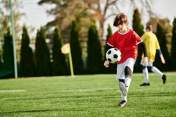 Un ragazzo in una maglia vibrante prende a calci un pallone da calcio su un campo verde sotto il sole luminoso. La sua espressione focalizzata mostra determinazione e passione per il gioco. — Foto stock